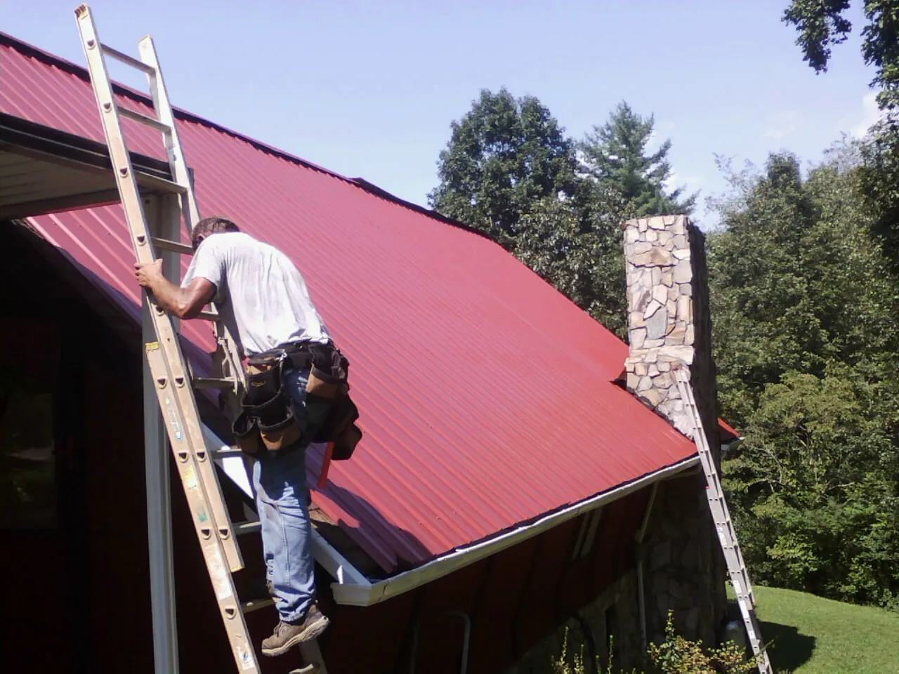Roofer on a ladder beside a red metal roof and stone chimney — Soffit & Fascia Repair by Roofing Brothers in Fort Worth